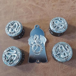 Könnte beinhalten: Eine Sammlung von silberfarbenen Metallknöpfen und einem Anhänger, alle mit Drachendesigns. Die Knöpfe haben schwarze Basen mit Zahlenmarkierungen. Der Anhänger ist ein drachenförmiger Charm mit einem Loch oben.