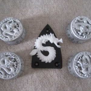 Peut inclure: Cinq boutons argentés avec un motif de dragon blanc et des chiffres noirs. Les boutons sont disposés en demi-cercle autour d'un pickguard de guitare noir avec un motif de dragon blanc.