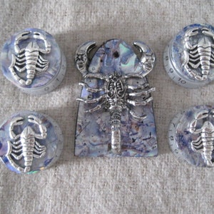 Puede incluir: Cinco pomos de escorpión plateados con fondos marmoleados azules y blancos. Los pomos están numerados del 1 al 10. El pomo central tiene un escorpión más grande con un fondo marmoleado azul y blanco.