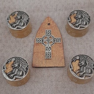 Puede incluir: Conjunto de cuatro perillas plateadas y doradas con temática de dragón y un colgante de cruz celta. Las perillas tienen un diseño de dragón plateado con detalles dorados. El colgante es de forma triangular marrón con una cruz celta plateada.