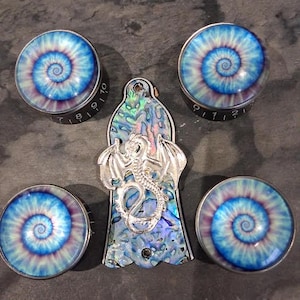 Könnte beinhalten: Eine Sammlung dekorativer Knöpfe und ein Anhänger. Die Knöpfe haben ein blaues, lila und weißes Spiraldesign. Der Anhänger ist ein silberner Drache auf einem blau, grün und lila schillernden Hintergrund.