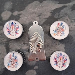 Puede incluir: Un conjunto de perillas decorativas y un colgante de metal. Las perillas presentan un diseño de atrapasueños con plumas coloridas y detalles florales. El colgante tiene un perfil nativo americano.