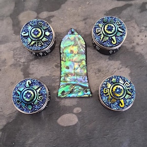 Puede incluir: Cuatro perillas decorativas y una pieza rectangular. Las perillas tienen un diseño azul, verde y amarillo con una base negra. La pieza rectangular es iridiscente con tonos verdes, azules y amarillos. Los artículos están sobre una superficie gris.