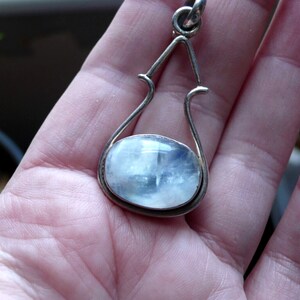 May include: A silver pendant featuring a large oval moonstone set within a delicate, ornate frame. The pendant is held in a hand.