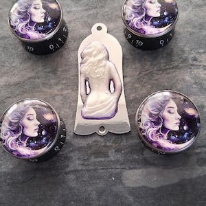 Könnte beinhalten: Eine Sammlung dekorativer Knöpfe und ein Anhänger mit einem Himmelsmotiv. Die Knöpfe haben einen schwarzen Sockel mit weißen Zahlen und eine Glasoberseite mit dem Profil einer Frau vor einem lila-blauen Galaxiehintergrund. Der Anhänger zeigt eine Frau.