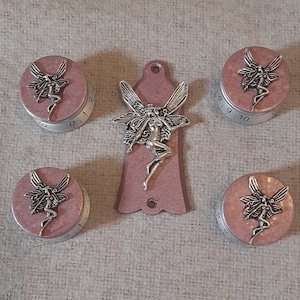 Könnte beinhalten: Satz von vier rosa und silbernen Knöpfen mit einem Feendesign und einem passenden rosa und silbernen Gitarrenplektrum mit einem Feendesign.