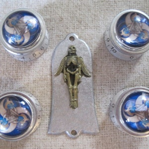 Könnte beinhalten: Vier silberne Knöpfe mit blauen und weißen wirbelnden Designs und schwarzen Zahlen. Eine silberne Metallplatte mit einer goldenen Skelettfigur.