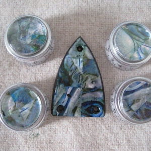 May include: Five silver knobs with blue and green marbled designs. The knobs have numbers printed on them. A blue and green marbled guitar pick is in the center.