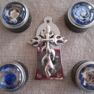 Könnte beinhalten: Ein silberner Gitarren-Pickguard mit einem Kreuz- und Flammen-Design. Der Pickguard ist von vier blauen und goldenen Gitarrenknöpfen umgeben.