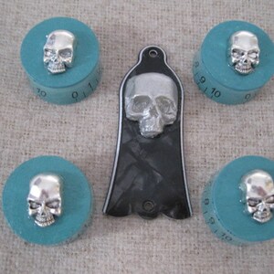 Puede incluir: Cinco pomos turquesa con detalles de calavera plateada y una placa de protecci&oacute;n de guitarra negra con un dise&ntilde;o de calavera plateada. Los pomos tienen los n&uacute;meros 7, 8, 9, 10 y 11 impresos en ellos.