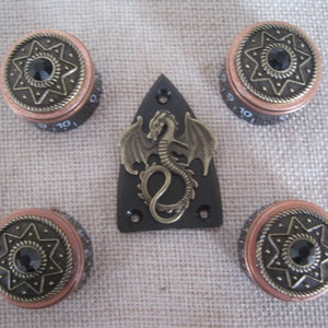 Puede incluir: Conjunto de cuatro botones de guitarra de cobre y latón con un diseño de estrella y una gema negra en el centro. Los botones están rodeados por una placa de protección negra con un diseño de dragón dorado.