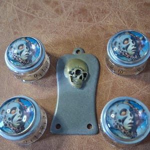 May include: A set of decorative knobs and a metal pendant with a skull design. The knobs have a face design with blue and teal accents, and a numbered dial. The pendant features a brass skull on a grey metal base.
