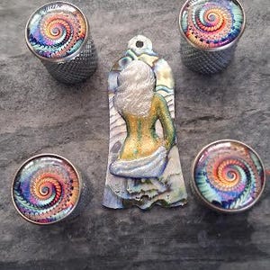 May include: An assortment of decorative items featuring a spiral design in vibrant colors. Includes four silver-toned thimbles and a pendant with a woman's figure. The thimbles and pendant are set against a textured gray surface.