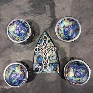 Op de afbeelding: Een assortiment handgemaakte sieradencomponenten. De afbeelding toont drie ronde, zilveromrande knoppen met wervelende blauwe, paarse en groene patronen, en een driehoekige hanger met een zilveren Keltisch kruisontwerp. De knoppen hebben zwarte bases met cijfers.