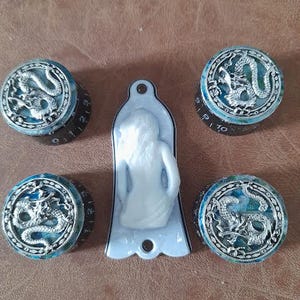 Op de afbeelding: Vijf decoratieve knoppen met een drakenontwerp en een wit beeldje. De knoppen hebben een blauw en zilver drakenontwerp op een zwarte basis. Het beeldje is wit en heeft een gebogen vorm.