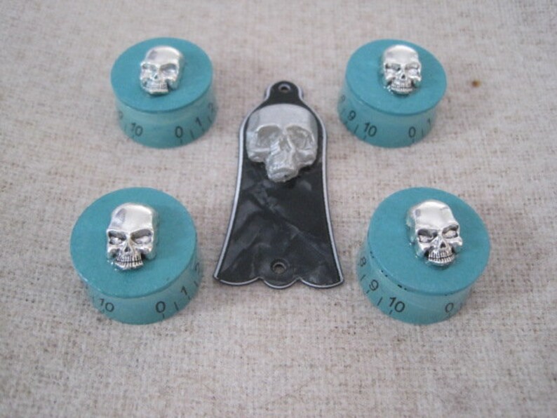 Puede incluir: Cinco pomos turquesa con detalles de calavera plateada y una placa de protecci&oacute;n de guitarra negra con una calavera plateada. Los pomos tienen los n&uacute;meros del 0 al 10 impresos.