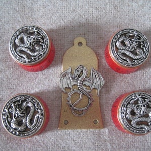 Puede incluir: Conjunto de cinco pomos rojos con diseños de dragones plateados. Los pomos son circulares y tienen un borde elevado. Los dragones están en un diseño circular con sus colas enroscadas alrededor de sus cuerpos. Los pomos son para una guitarra u otro instrumento musical.