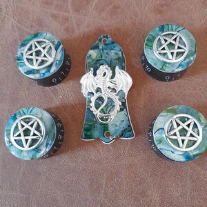 Könnte beinhalten: Satz von fünf Gitarren-Stimmknöpfen mit Pentagramm-Design und einem passenden Gitarren-Plektrum mit Drachen-Design. Die Knöpfe sind schwarz mit weißen Zahlen und das Plektrum ist blau und grün mit einem silbernen Drachen.