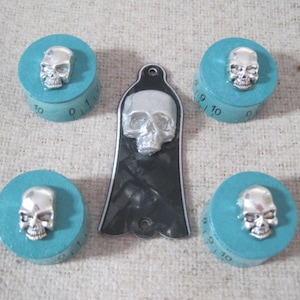 Puede incluir: Conjunto de cuatro pomos turquesa con detalles de calavera plateada. Cada pomo tiene una base circular con los números del 0 al 11 impresos alrededor del borde. Los pomos están dispuestos en forma de cuadrado con una púa de guitarra de calavera negra y blanca en el centro.