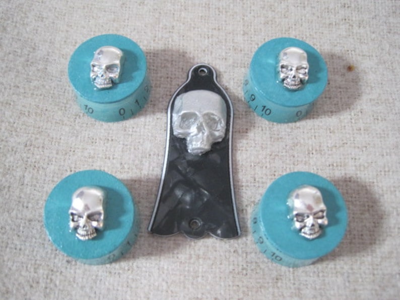 Puede incluir: Conjunto de cuatro pomos turquesa con detalles de calavera plateada. Cada pomo tiene una base circular con los n&uacute;meros del 0 al 11 impresos alrededor del borde. Los pomos est&aacute;n dispuestos en forma de cuadrado con una p&uacute;a de guitarra de calavera negra y blanca en el centro.