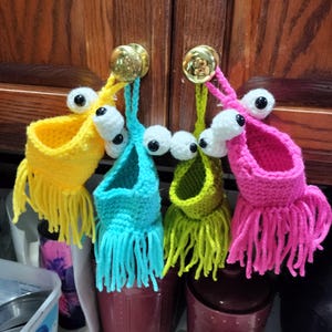 May include: Four crocheted hanging pockets with googly eyes and fringe. The pockets are yellow, blue, green, and pink.