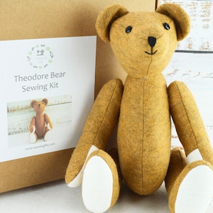 May include: A brown felt teddy bear sewing kit with white felt paws. The kit includes all the materials needed to sew a teddy bear. The kit is packaged in a brown cardboard box with a white label that says "Theodore Bear Sewing Kit".