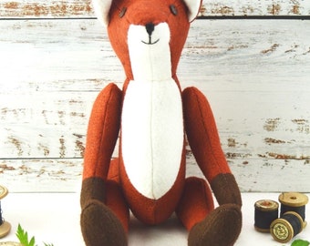 May include: A handmade plush fox toy with orange, white, and brown felt. The fox has a white chest and inner ears, with brown feet and a stitched black nose and eyes. The toy is sitting on a white surface.