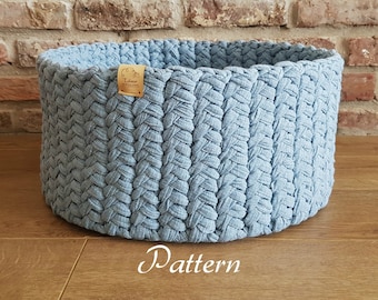 May include: A light blue crocheted storage basket with a woven pattern. The basket is sitting on a wooden floor in front of a brick wall.