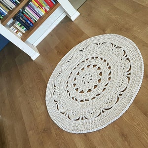 May include: A white crocheted round rug with a delicate lace pattern. The rug is on a light brown wood floor.