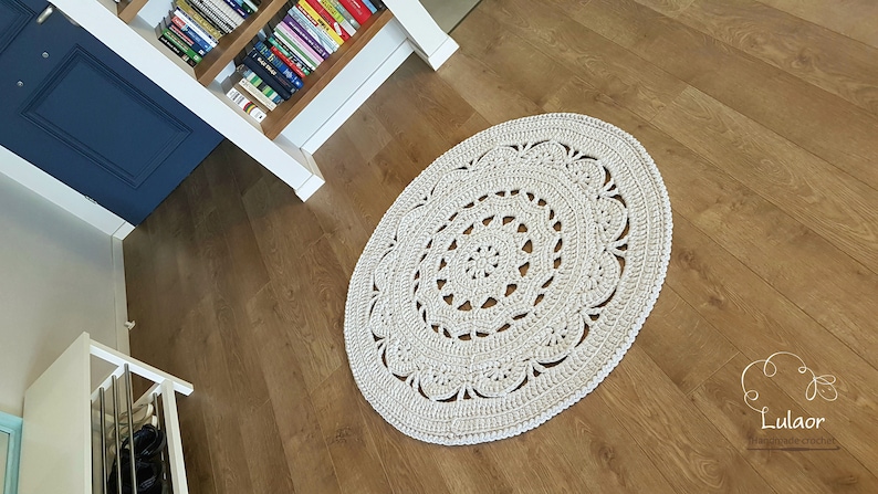 May include: A white crocheted round rug with a delicate lace pattern. The rug is on a light brown wood floor.