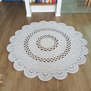 May include: A round, light gray crocheted rug with a scalloped edge. The rug features a detailed openwork design in the center and along the border. The rug is placed on a wooden floor.