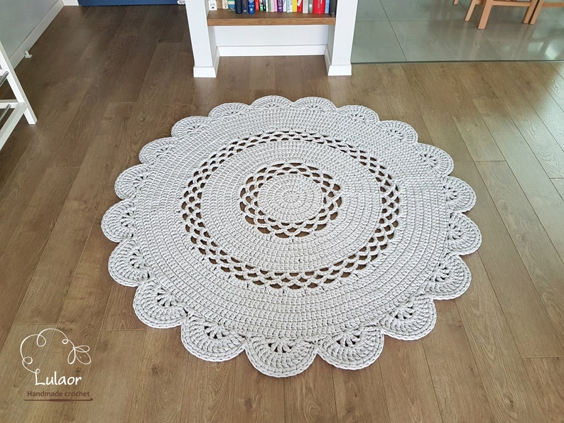 May include: A round, light gray crocheted rug with a scalloped edge. The rug features a detailed openwork design in the center and along the border. The rug is placed on a wooden floor.