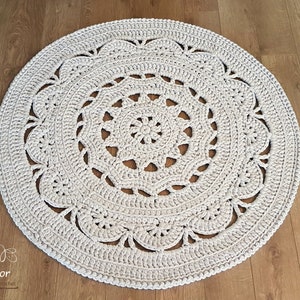 May include: A white crocheted round rug with a delicate lace pattern. The rug has a circular design with a central motif and a border of intricate crocheted details.