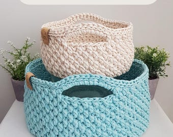 May include: Two round, hand-crocheted baskets with integrated handles, one in cream and one in light blue. Each basket features a textured stitch pattern and a small brown leather-like tag on the side. Text reads: 2 SIZES • PDF PATTERN.