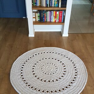 May include: A large, round, cream-colored crocheted rug with a circular pattern. The rug is on a wooden floor.