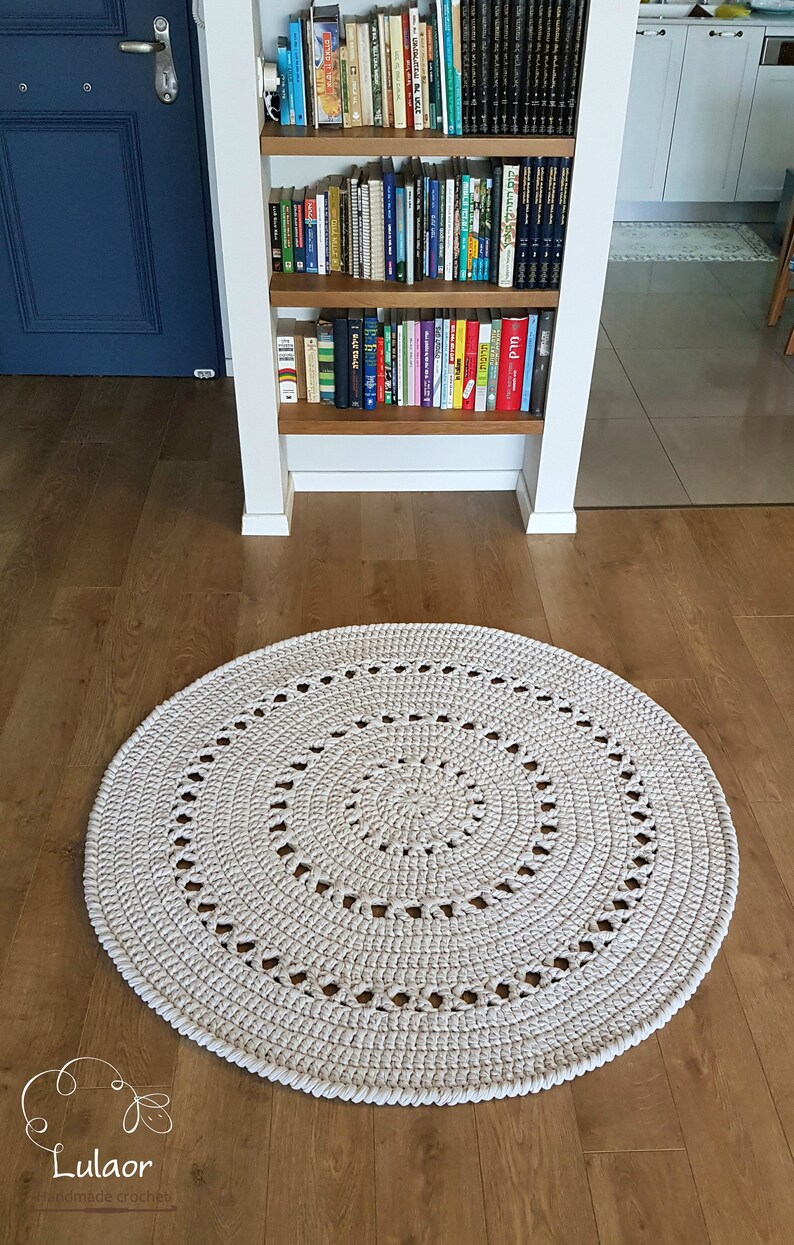 May include: A large, round, cream-colored crocheted rug with a circular pattern. The rug is on a wooden floor.