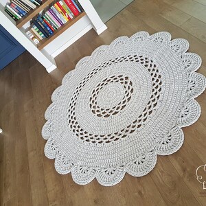 May include: A light gray, handmade, oval crochet rug with a scalloped edge. The rug features concentric circles of crochet work, creating a textured surface. The rug is placed on a wooden floor.