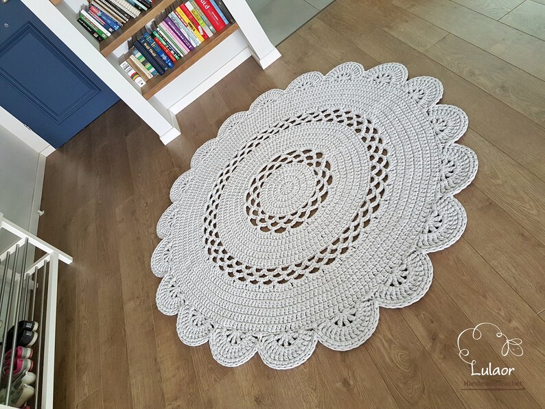 May include: A light gray, handmade, oval crochet rug with a scalloped edge. The rug features concentric circles of crochet work, creating a textured surface. The rug is placed on a wooden floor.