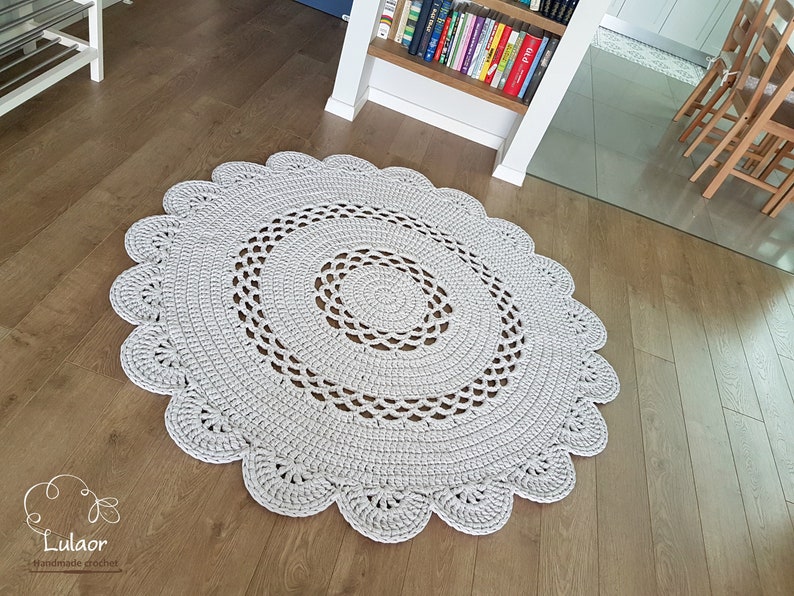May include: A light gray, round, crocheted rug with a scalloped edge. The rug features a spiral design in the center and is made of a soft, textured yarn. The rug is approximately 47 inches in diameter, perfect for a living room or bedroom.