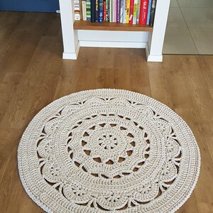 May include: A large, round, crocheted rug in a cream colour with a detailed, intricate design. The rug is made with thick yarn and has a delicate, lacy pattern.