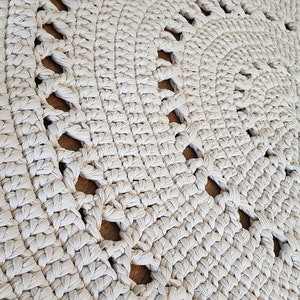 May include: A close-up of a cream-colored crocheted rug with a circular design and openwork details. The rug is made with thick yarn and has a textured surface.