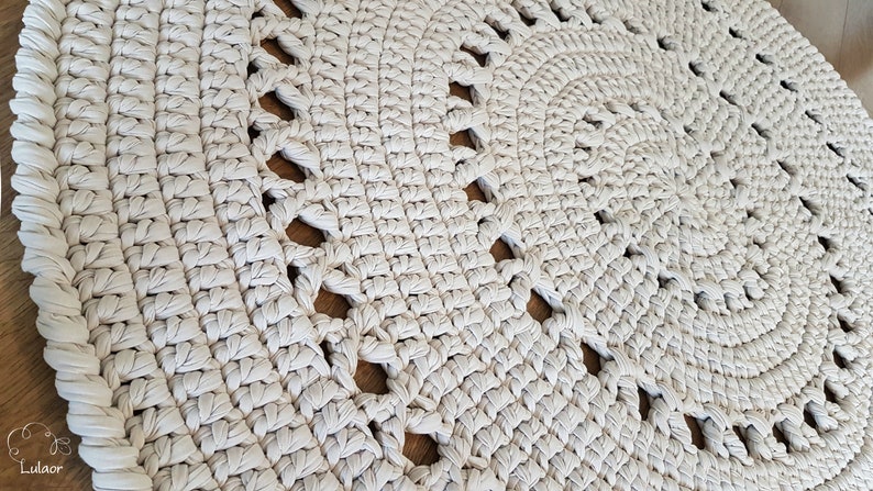May include: A close-up of a cream-colored crocheted rug with a circular design and openwork details. The rug is made with thick yarn and has a textured surface.