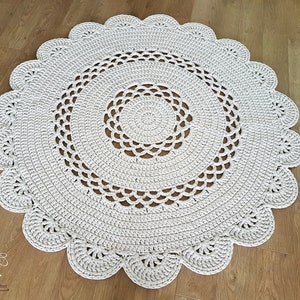 May include: A round, off-white crochet rug with a scalloped edge. The rug features concentric circles of openwork and solid crochet stitches. The rug is approximately 47 inches in diameter, suitable for a living room or bedroom.
