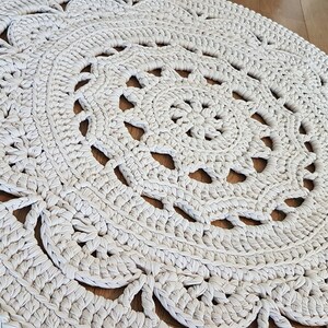 May include: A white crocheted round rug with a delicate floral pattern. The rug has a circular design with intricate details and a soft, textured surface.