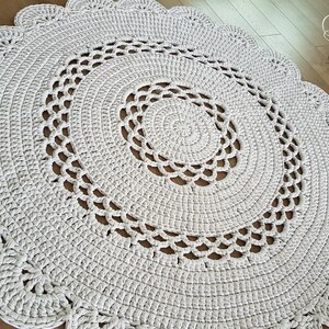 May include: A round, off-white crochet rug with a scalloped edge. The rug features a spiral design in the center and decorative openwork patterns. The handmade rug is a neutral color and would complement any room.