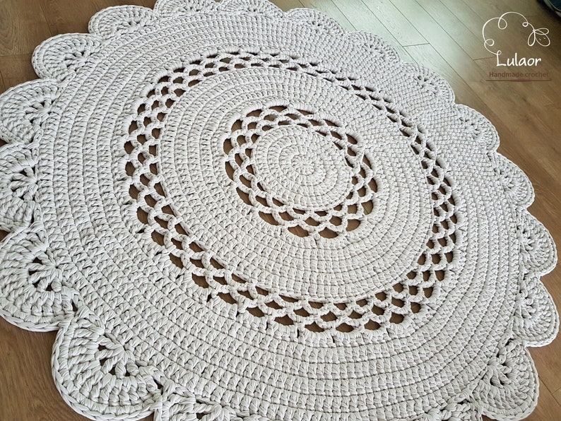 May include: A round, off-white crochet rug with a scalloped edge. The rug features a spiral design in the center and decorative openwork patterns. The handmade rug is a neutral color and would complement any room.