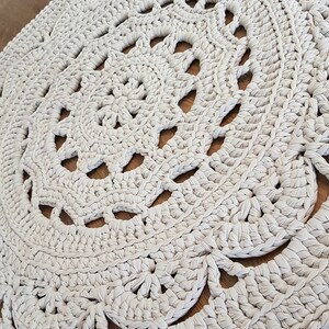 May include: A large, round, crocheted rug in a cream colour with a delicate, lacy pattern. The rug is made with thick yarn and has a soft, textured surface.