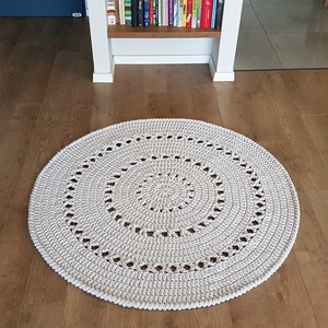 May include: A white crocheted round rug with a circular pattern in the center. The rug is on a wooden floor.