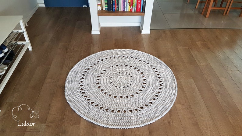 May include: A white crocheted round rug with a circular pattern in the center. The rug is on a wooden floor.