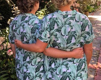 May include: Two people wearing matching green and white tropical print pajamas with short sleeves and shorts. The pajamas have a white trim around the collar and sleeves.